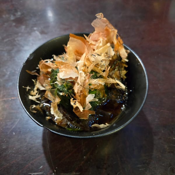 A small black bowl of green seaweed salad topped with light brown bonito flakes.