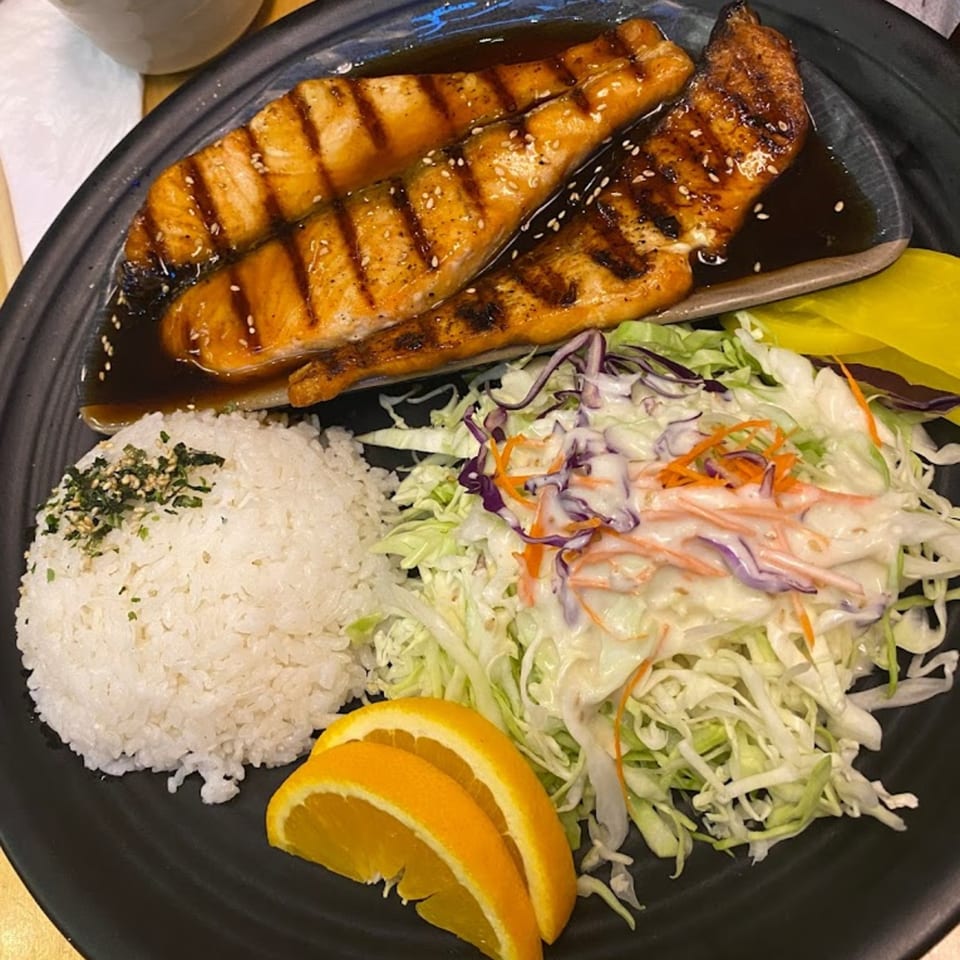 Grilled salmon with teriyaki sauce, white rice, coleslaw, and orange slices on a black plate.